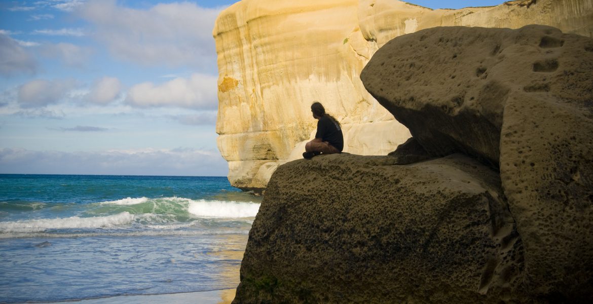 A man sits on a large boulder at Dunedin, New Zealand's Tunnel Beach. The sun illuminates a large cliff in the distance as blue and green waves crash on the shore.