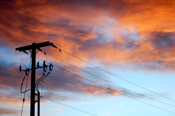 The sun sets behind a power pole along Interstate 86, Eastern Idaho. Pink, orange, and purple clouds hover over the silhouetted power pole and power lines.