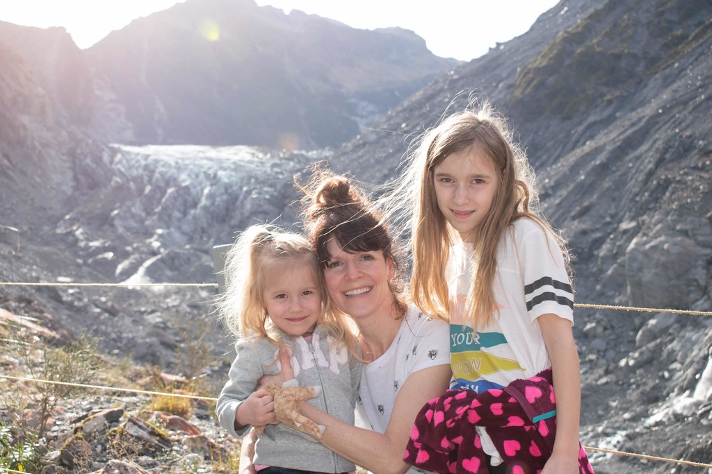 A woman and two young girls sit in the foreground with a distant view of Fox Glacier behind them. The receding Fox Glacier is visible from the glacier viewing area. Stone debris and slate are visible in much of the foreground, with only a small edge of the glacier still visible at the top of the valley.