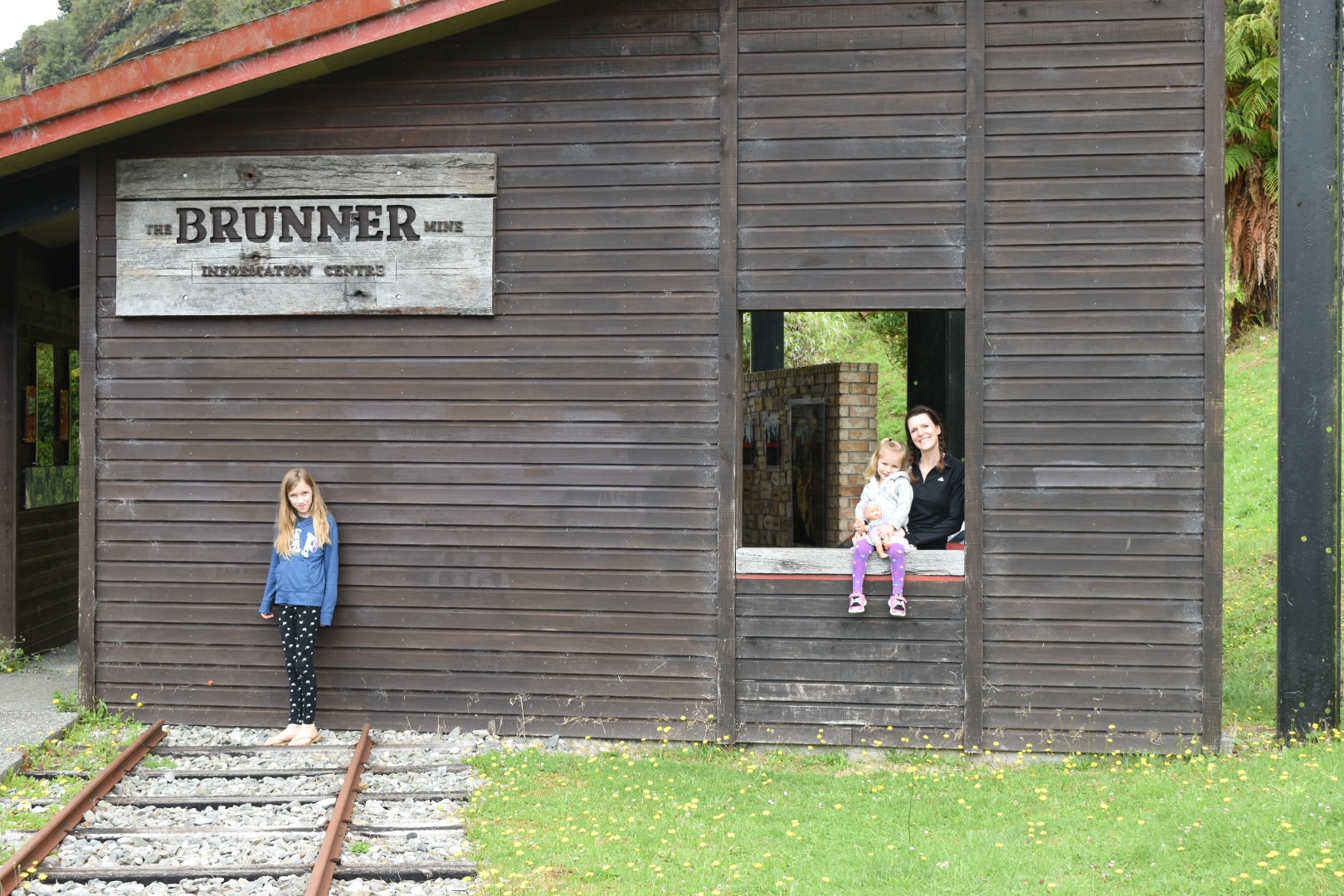 A mother and daughter pose in an open window at the Brunner, New Zealand, while another daughter stands nearby, on railroad tracks that lead toward a museum building on the site.