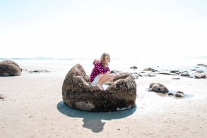 A young girl crouches inside a massive Moeraki Boulder sitting on the sandy Pacific Coast of New Zealand. The top of the boulder has eroded, leaving a large, concave opening in which the girl sits. The Pacific Ocean is visible off New Zealand's South Island on the East Coast near the village of Moeraki.