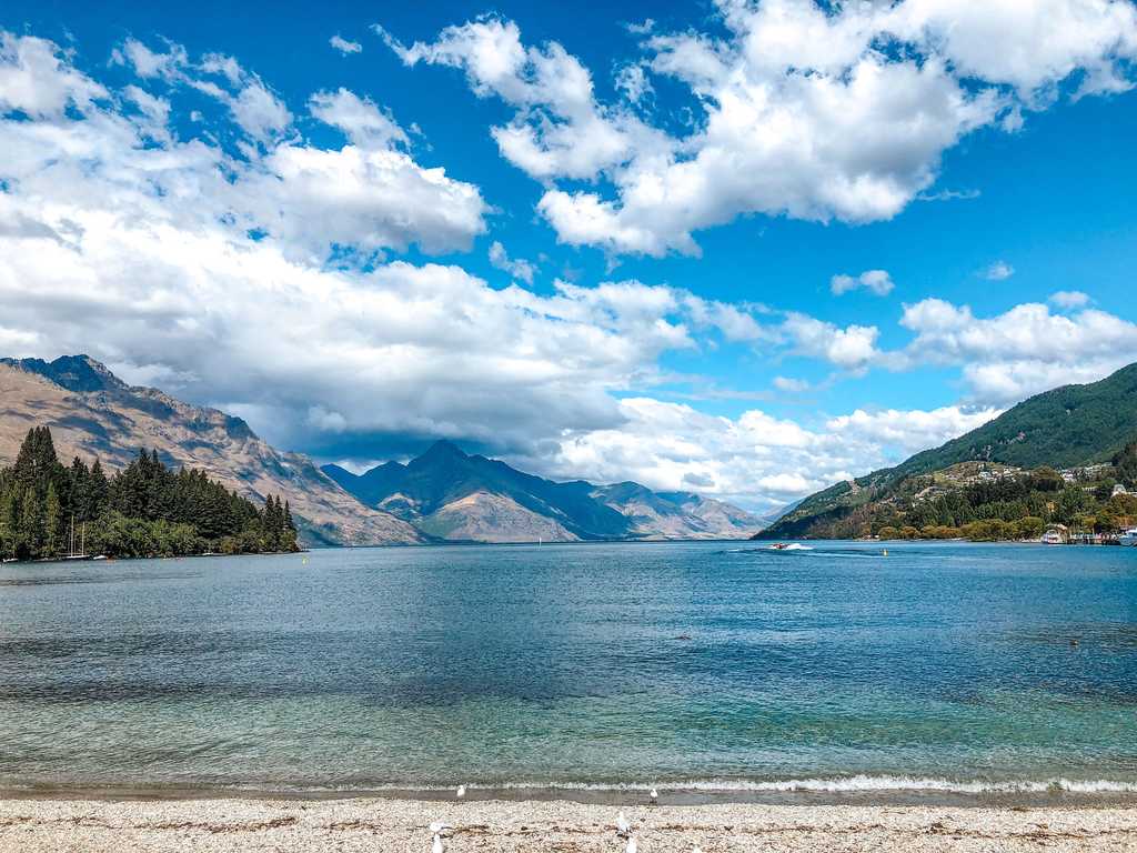 A view of Lake Wakatipu from Queenstown's waterfront park. Bayonet Peaks and Walter Peak are both visible over the lake. Blue-green, turquoise water sits beneath a vibrant blue sky, as fluffy white clouds float by.