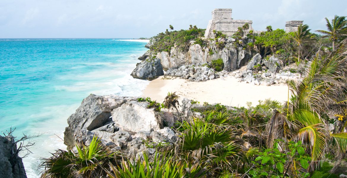 A view of the main stone structure at the Pre-Colombian Mayan ruins at Tulum, Yucatan Peninsula, Mexico. The stone structure sits on a cliff overlooking the turquoise-blue waters of the Caribbean Sea. A white sand beach, rock outcroppings, and dark green foliage fill the foreground.