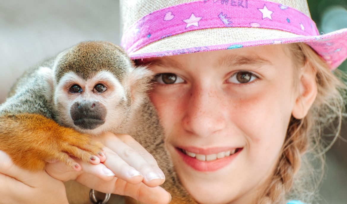 A young girl smiles as she holds a small monkey on her shoulder while wearing a pink-brimmed straw hat. The monkey is part of the various tourist attractions at the Pre-Colombian Mayan ruins of Tulum, Mexico, overlooking the Caribbean Sea on the Yucatan Peninsula.