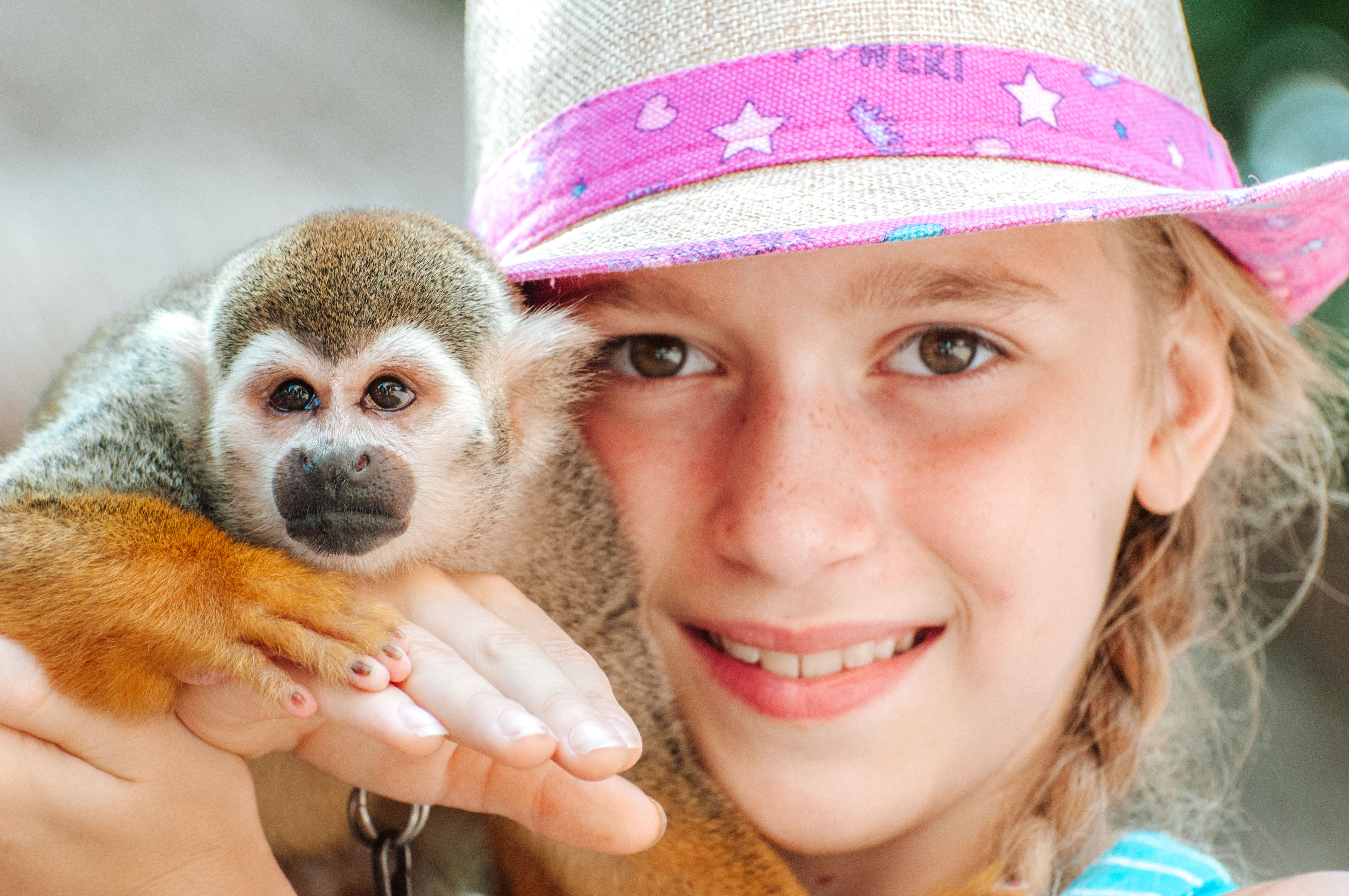 A young girl smiles as she holds a small monkey on her shoulder while wearing a pink-brimmed straw hat. The monkey is part of the various tourist attractions at the Pre-Colombian Mayan ruins of Tulum, Mexico, overlooking the Caribbean Sea on the Yucatan Peninsula.