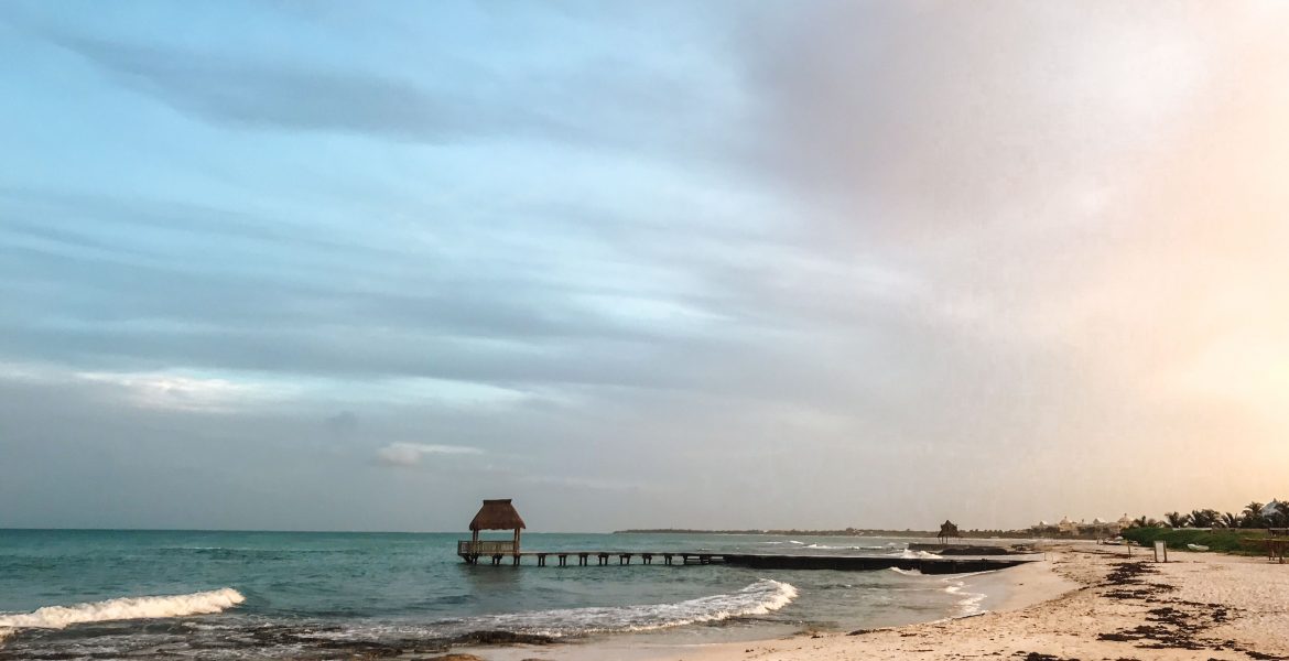 A small pier juts out into the Caribbean Sea between Mexico's Playa de Carmen and Cancún, at the Mayan Palace and Grand Mayan luxury resorts along the Riviera Maya. Clouds reflect the setting sun over the turquoise-blue water and white sand.