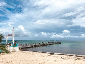 A small pier juts out into the Caribbean Sea in Mexico's Puerto Morelos, along the Riviera Maya. Clouds float over turquoise-blue water and white sand.