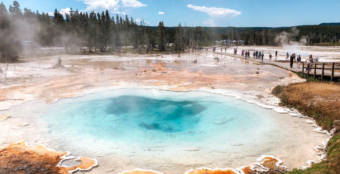 A large thermal spring connected to Celestine hot spring near Fountain Paint Pots in Yellowstone National Park, Wyoming. Deep blue sky contrasts with white clouds over the stark bacteria mats and mineral earth near the spring. The boiling water creates a deep blue and turquoise hue, surrounded by orange, yellow, and brown bacteria mats and a wide walkway for tourists to walk to viewing platforms.