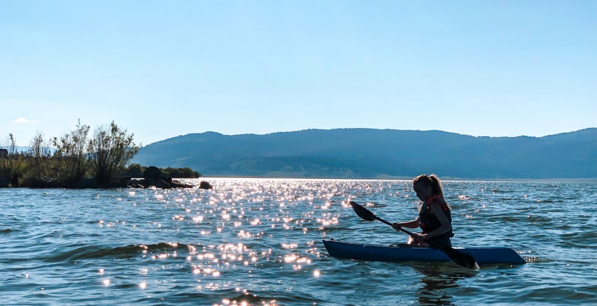 A young girl paddles her kayak across Henrys Lake in Northeastern Idaho, near Yellowstone National Park. She is silhouetted against the deep, blue water, and the sun shimmers and sparkles across the water. Blue sky and mountains can be seen in the distance, the edge of the Targhee National Forest.