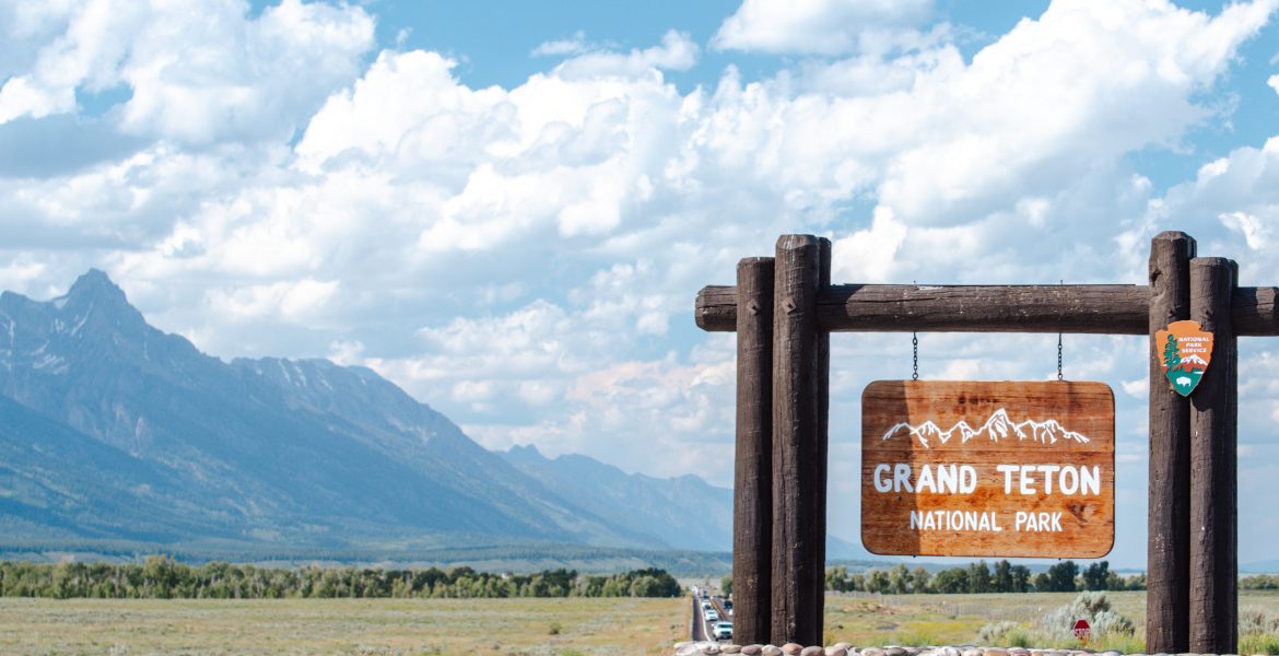A view of the National Park Service sign at the entrance to Grand Teton National Park, Wyoming, just outside of Jackson, Wyoming. The Tetons sit beneath white clouds and a blue summer sky.